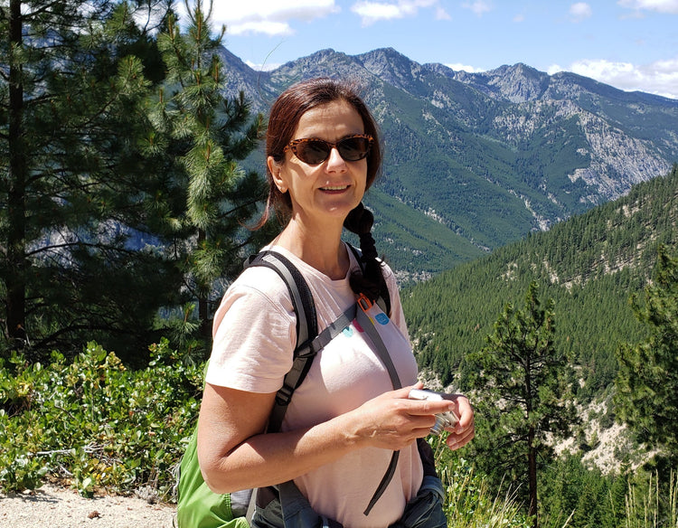A woman wearing sunglasses, a light pink t-shirt, and a green backpack stands outdoors in a mountain landscape with pine trees and rocky peaks in the background.